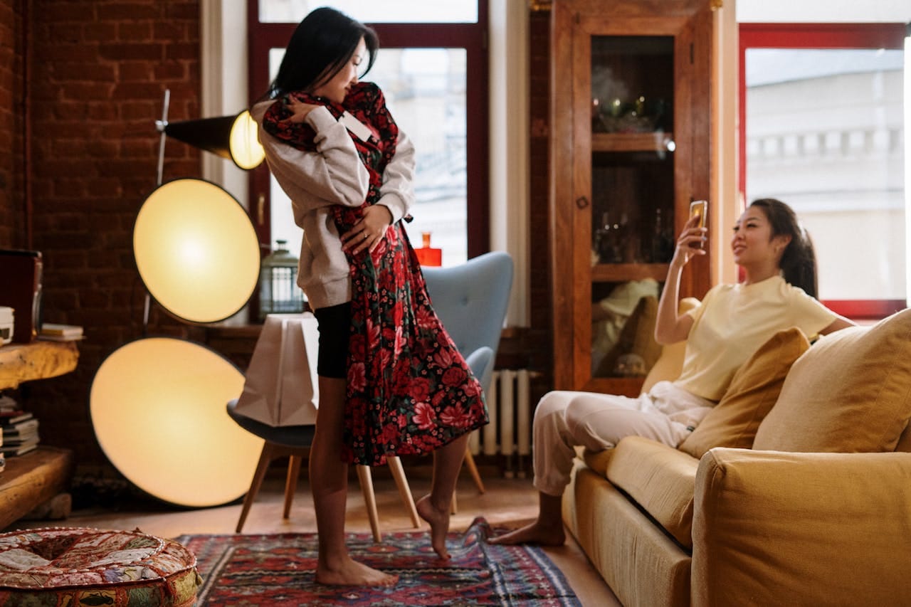 Two friends relaxing indoors, one fitting a dress while the other uses a smartphone.